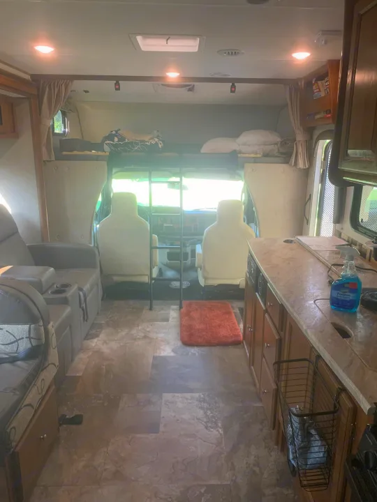 RV rental interior: A motorhome's compact living space, featuring a kitchen counter, front cockpit, and overhead bunk bed.. Forest River Coachmen Leprechaun 2018