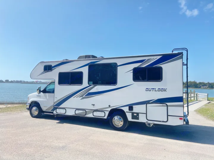White rv rental with blue/grey accents parked by calm water under a clear blue sky, ideal for adventures.. Winnebago Outlook 2020