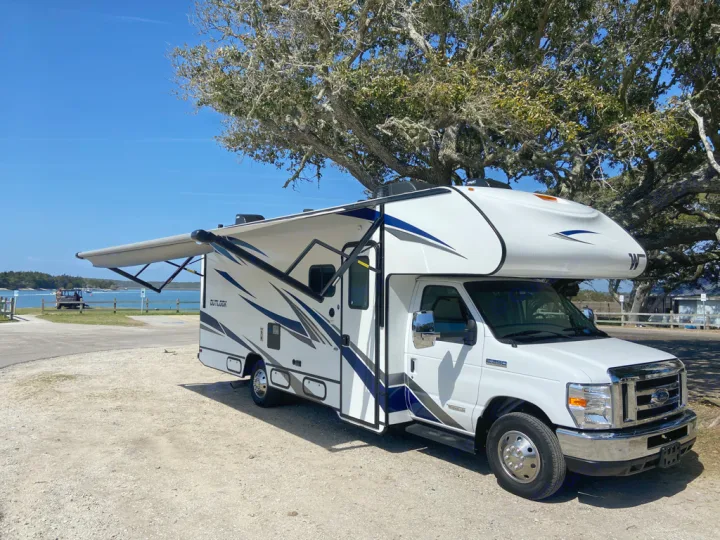 Enjoying the outdoors! This white and blue Class C RV rental with extended awning is parked by a scenic bay under a clear sky.. Winnebago Outlook 2020