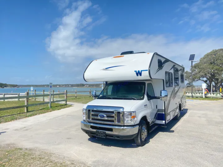 White RV rental parked by a scenic waterfront on a sunny day, ready for your next journey.. Winnebago Outlook 2020