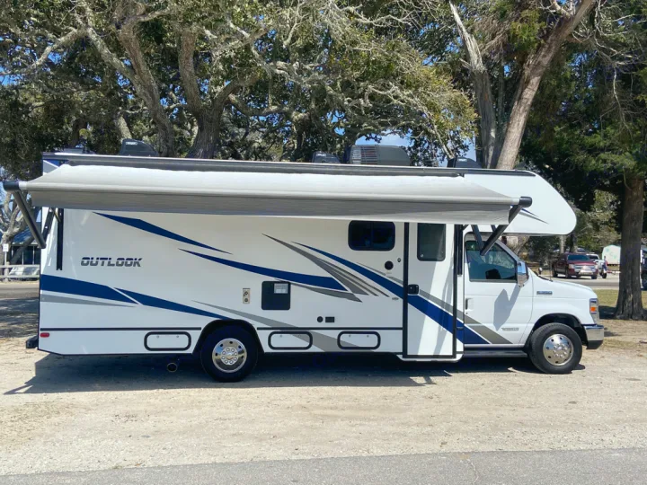 White Class C RV rental with an extended awning, parked outdoors among trees. Perfect for your next road trip or camping.. Winnebago Outlook 2020