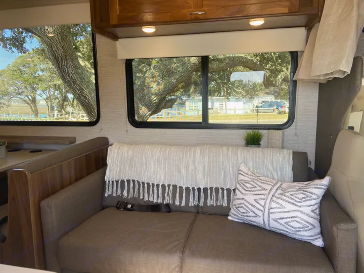 RV rental living area with a sofa, throw blanket, dinette, and scenic views of trees and water outside.. Winnebago Outlook 2020