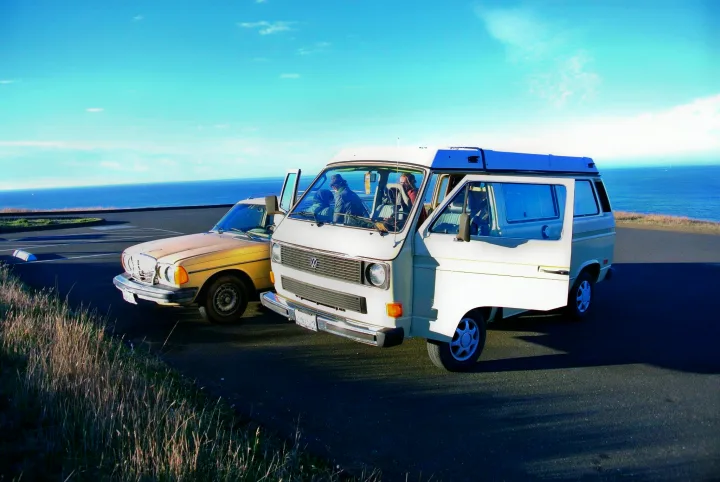 Vintage VW camper van and sedan by the bright blue ocean, great for an RV rental road trip.. Volkswagen Vanagon 1984