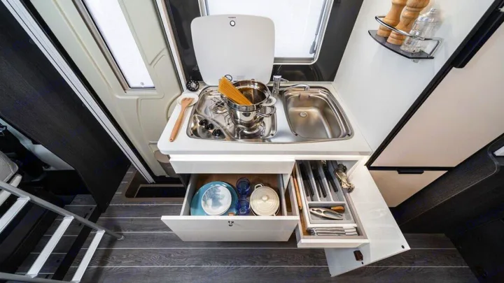 A compact RV rental kitchen with a sink, stovetop, and open drawers showing organized dishes and utensils.. Roller Team Kronos 271 M 2022