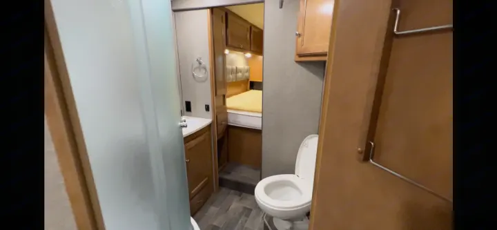 This RV rental features a compact bathroom with a frosted shower on the left, a sink, and a toilet on the right, connected to a bedroom.. Winnebago Vista 2019