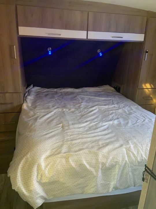 An RV rental bedroom features a bed with white patterned bedding, built-in wooden cabinets, and blue headboard lights.. Grand Design 2800BH 2021