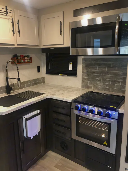 Modern RV rental kitchen featuring stainless steel microwave, oven, cooktop, sink, light countertops, and dark cabinets.. Crossroads Cruiser Aire 2021