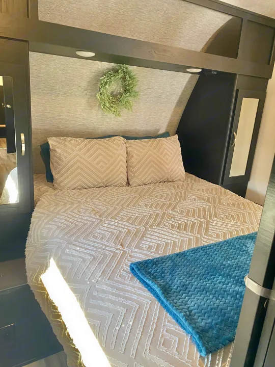Cozy RV rental bedroom featuring textured beige bedding, a folded blue blanket, built-in storage, and overhead lighting.. Coachmen Freedom express select 2022