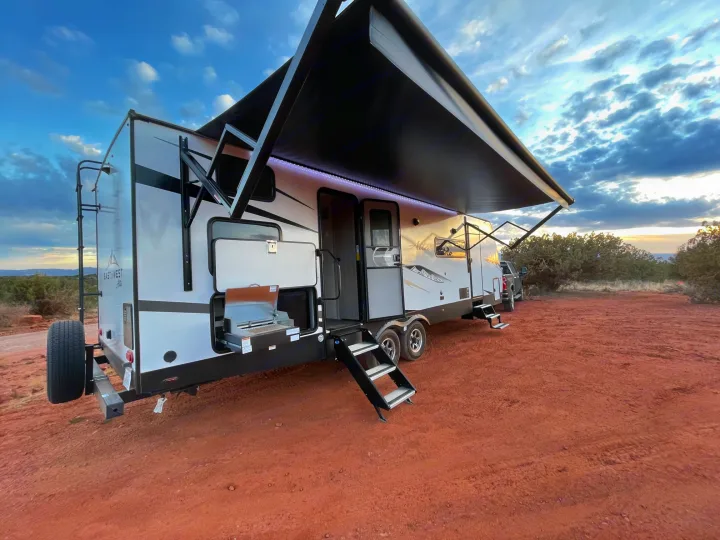 This RV rental travel trailer features an extended awning, outdoor kitchen, and steps, parked on vibrant red dirt.. East to West Alta 2022