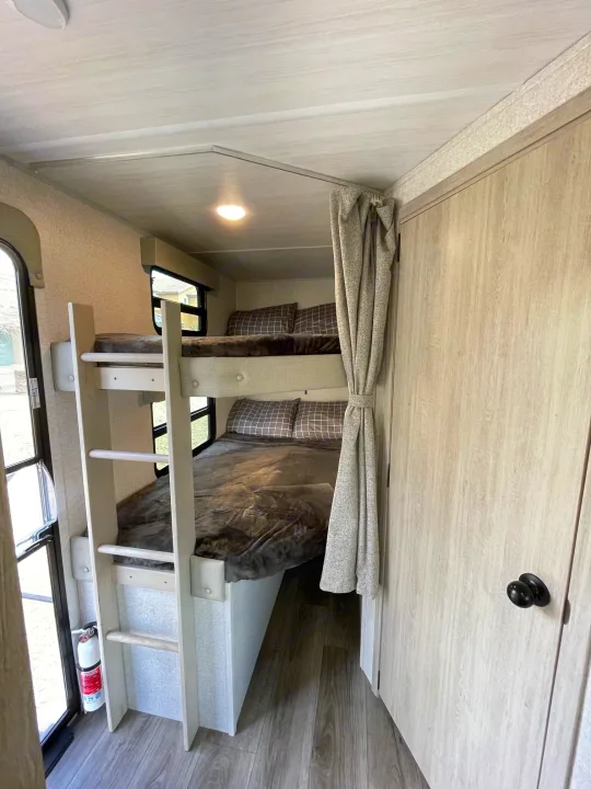 Inside an RV rental, showing light-colored bunk beds with a ladder, privacy curtain, and fire extinguisher below.. East to West Alta 2022