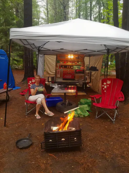 Relaxing camping scene with a woman by the fire, a teardrop RV rental, and a tent under a canopy.. The Teardrop Trailer Diamond in the rough 2021