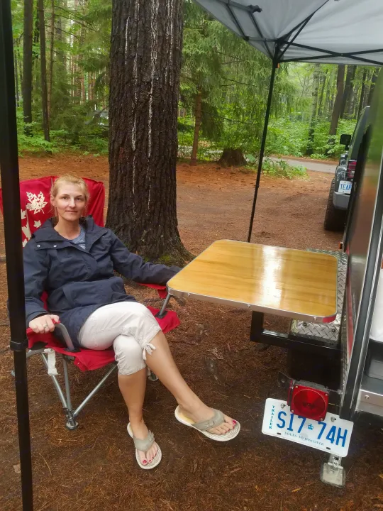Woman seated at her outdoor table attached to an RV rental trailer. Perfect for relaxing at a woodsy campsite.. The Teardrop Trailer Diamond in the rough 2021