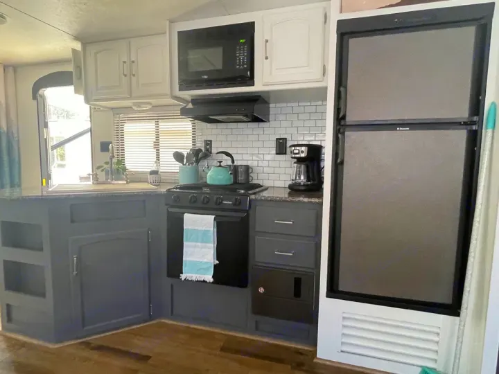 Renovated RV rental kitchen showing compact stove, sink, fridge, white upper & dark gray lower cabinets, and subway tile.. Keystone RV Springdale 2015