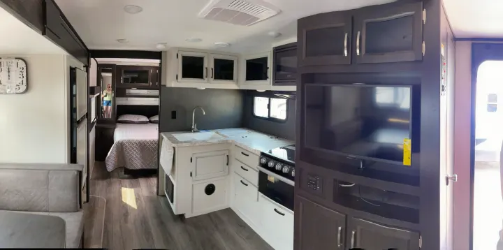 Inside a bright, modern RV rental: a wide view of the interior shows a kitchen, a bedroom with a bed, and living area with dinette and TV.. Jayco Jay Feather 2021