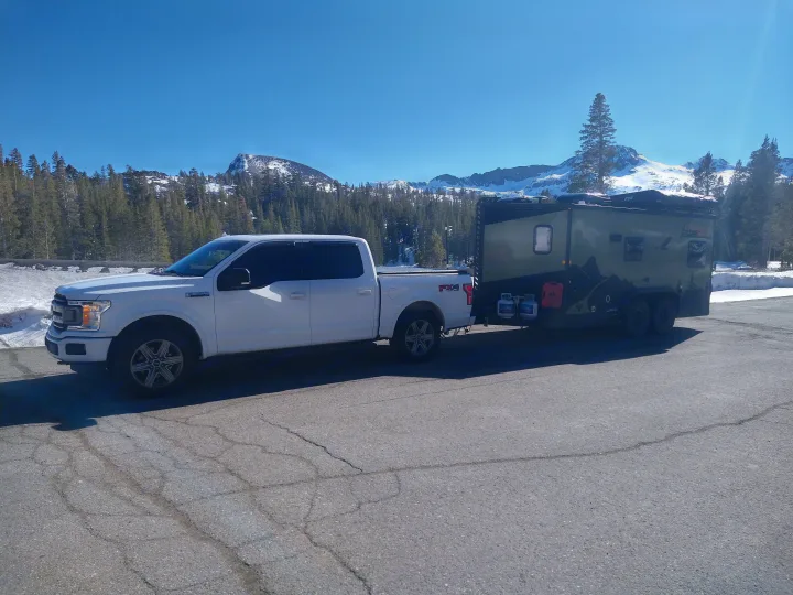 White truck towing a rugged RV rental camper with propane tanks, set against majestic snowy mountains and a forest.. Xplorer X22 2022