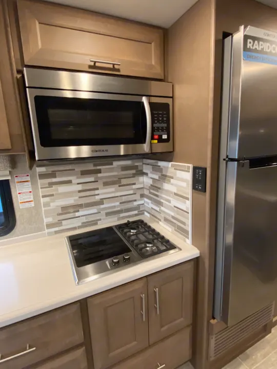 Inside an RV rental: a compact kitchen with a microwave, gas cooktop, sleek cabinets, and stainless steel fridge.. Thor 37.1 2022