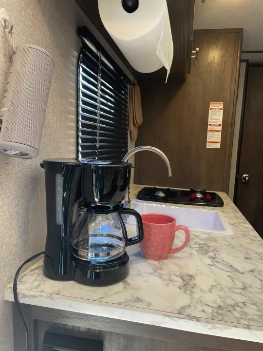 Coffee setup in an RV rental kitchen: a black coffee maker, pink heart mug, and sink on a marble counter. Blinds, speaker & paper towels visible.. Dutchmen Coleman Lantern 2022