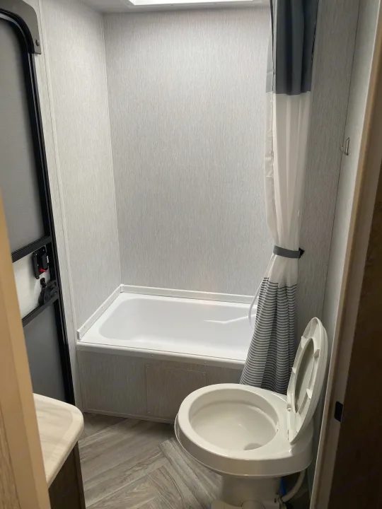 This RV rental bathroom features a compact tub/shower, open toilet, and gray striped curtain, with textured walls and floor.. Forest River 263BHXL 2022