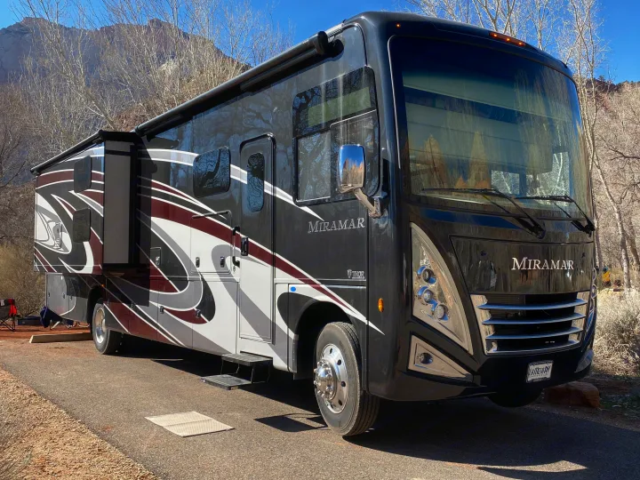 Black Thor Miramar RV rental with an extended slide-out, parked on asphalt at a sunny campground surrounded by trees & mountains.. Thor 37.1 2022