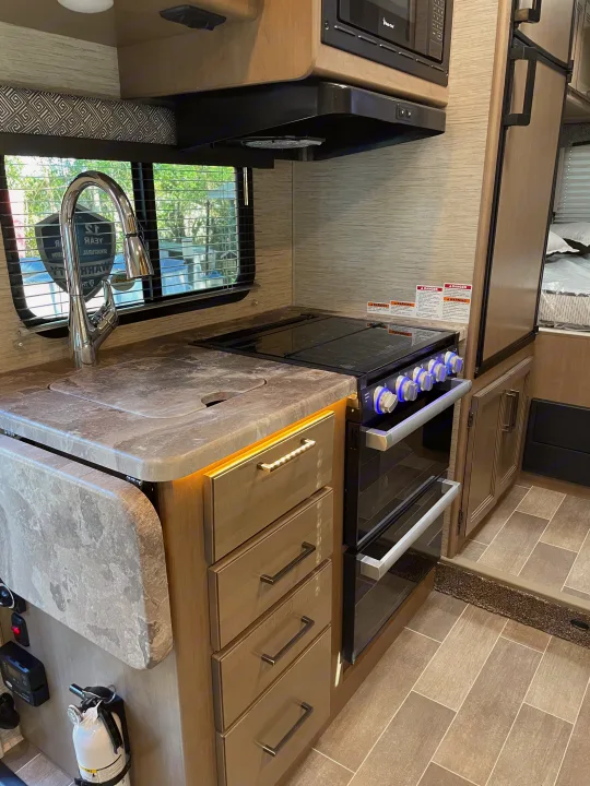 An RV rental kitchen with a sink, stovetop, oven, microwave, and light wood cabinets, offering efficient cooking and living space.. Thor Magnitude GA22 2022
