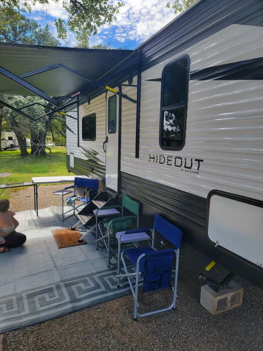 Campsite setup: A white RV rental with an awning, chairs, and outdoor mat on gravel. Relaxing outdoor scene.. Keystone Hideout 2021