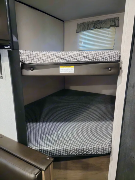 An RV rental bunk bed with safety netting and window. A caution label for elevated beds and a Roku TV are visible.. Keystone Hideout 2021