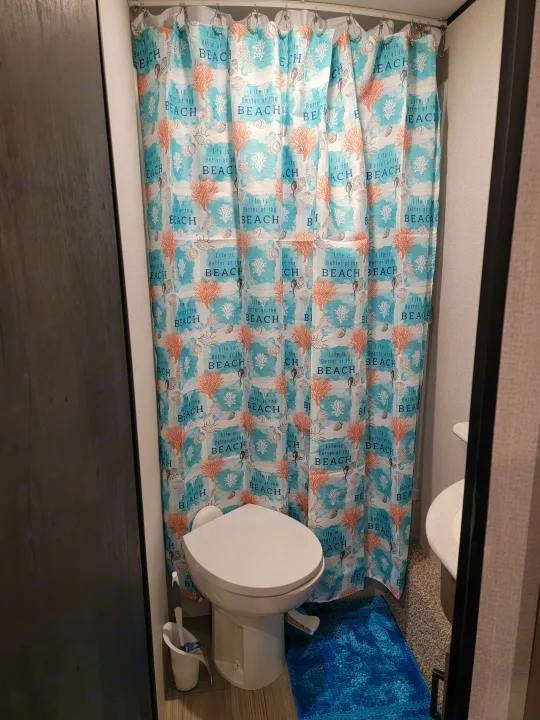 RV rental bathroom featuring a beach-themed shower curtain, toilet, and blue bath mat.. Keystone Hideout 2021