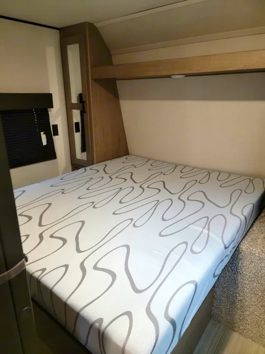 Interior view of an RV rental bedroom, featuring a patterned mattress, wooden nightstand with mirrored storage, and floor.. Keystone Hideout 2021