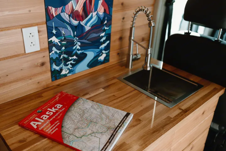**RV rental** interior: a cozy wooden kitchenette with a sink, an Alaska map book, and nature-themed wall art for travel planning.. Dodge Ram 2500 (Class B) 2021