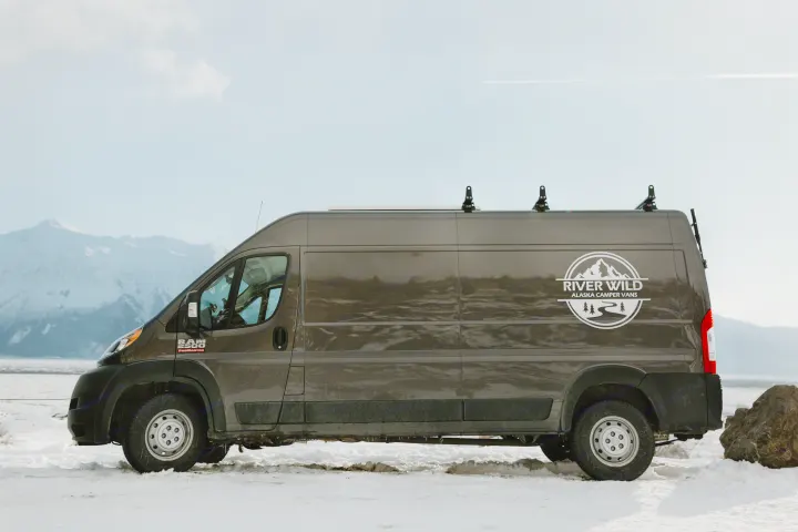 River Wild Alaska Camper Vans' RV rental, a grey Ram ProMaster, parked in a snowy landscape with mountains.. Dodge Ram 2500 (Class B) 2021
