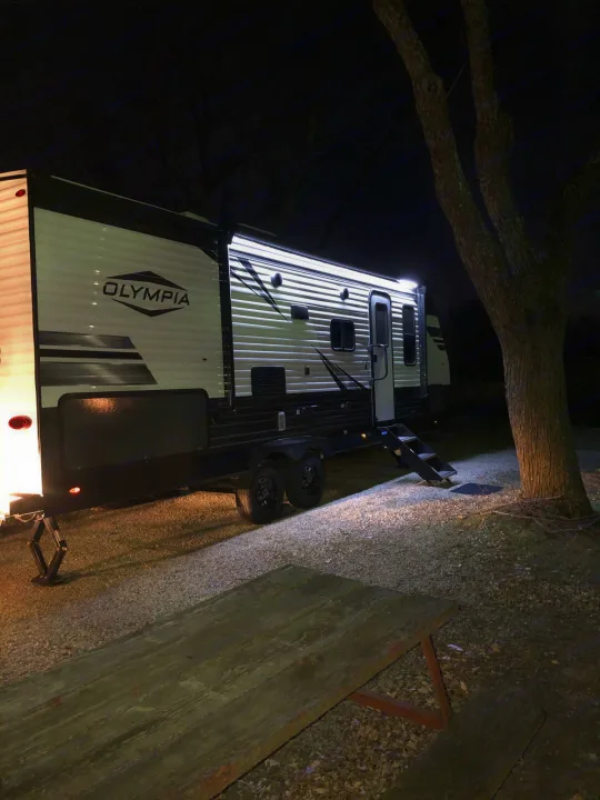 An Olympia travel trailer, lit up at night, parked at a campsite next to a picnic table. Perfect for an RV rental adventure!. Highland Ridge Olympia 26BHS 2022