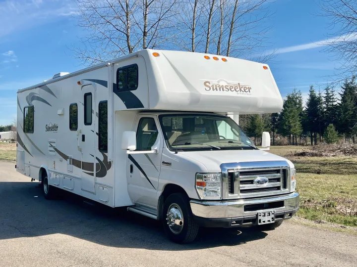 This white Sunseeker Class C rv rental, a perfect motorhome for travel, is parked on a paved lot under a sunny blue sky with bare trees.. Forest River Sunseeker 2009