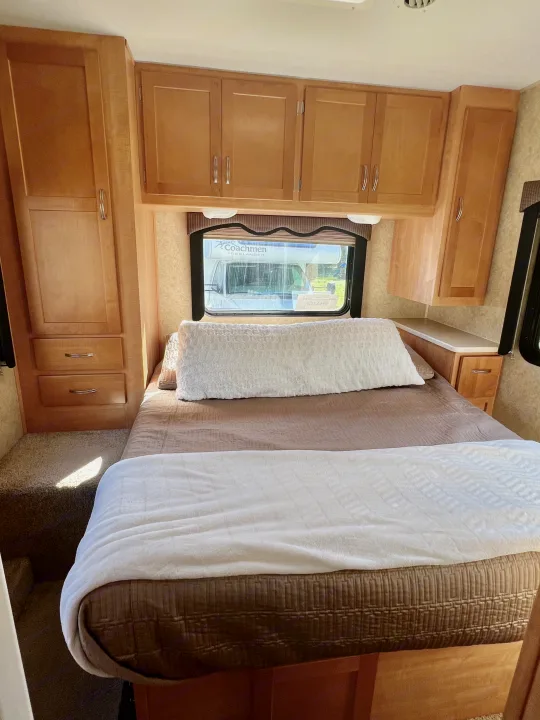 Cozy rv rental bedroom: light wood cabinets, comfy bed with white and brown bedding, and a window looking out.. Forest River Sunseeker 2009