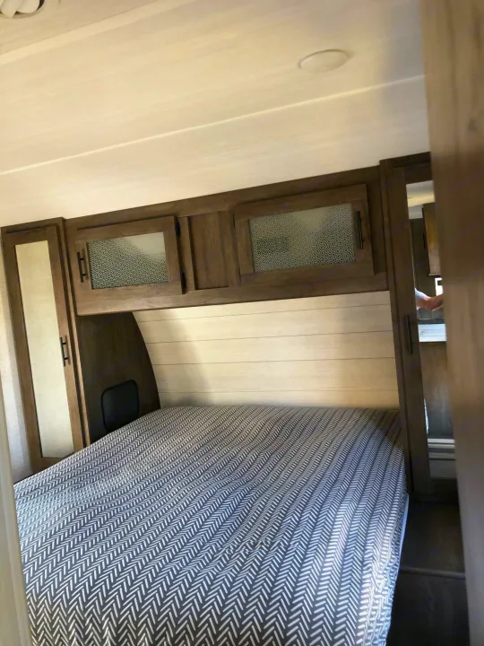 RV rental bedroom: a cozy space with a herringbone-patterned bed, overhead wood cabinets, and light shiplap walls.. Forest River Wildwood 2020