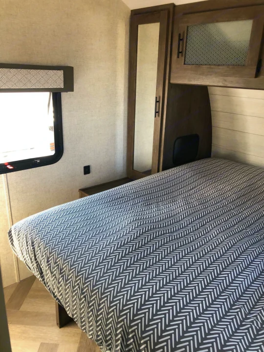 RV rental bedroom showing a patterned bed, mirrored cabinet, window, and modern finishes.. Forest River Wildwood 2020