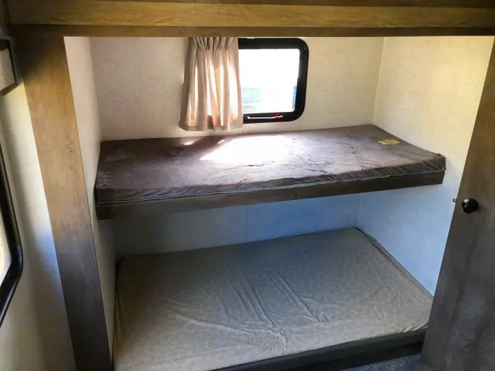 Inside an RV rental, showing a set of bunk beds with mattresses. The upper bunk features a window with a curtain.. Forest River Wildwood 2020