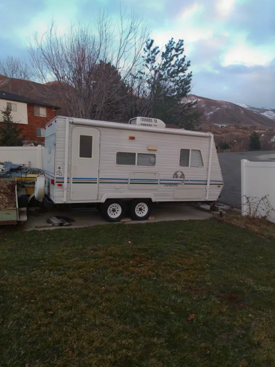 A white travel trailer, possibly an RV rental, parked on a paved driveway in a yard with mountains and houses in the background.. Sportsmen Coyote 2002