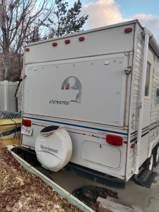 Rear of a white "Coyote" RV rental travel trailer with spare tire & Utah license plate, parked outdoors.. Sportsmen Coyote 2002
