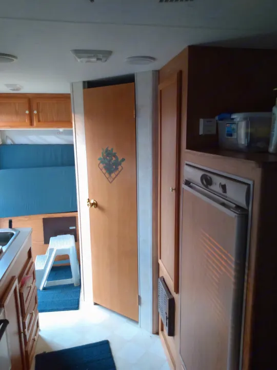 An RV rental interior features a functional kitchen, compact bed, refrigerator, and wooden cabinetry, creating a cozy mobile living space.. Sportsmen Coyote 2002