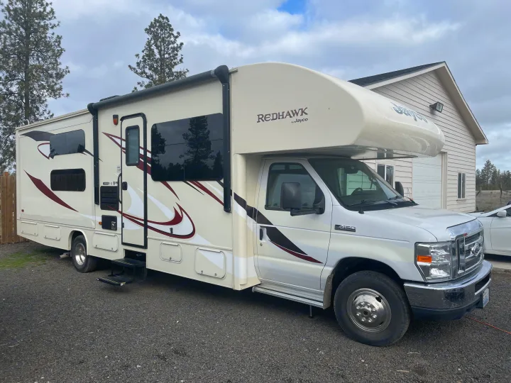 A white Jayco Redhawk Class C motorhome, an ideal RV rental, parked on a gravel surface near a beige garage under a cloudy sky.. Jayco Redhawk 2016