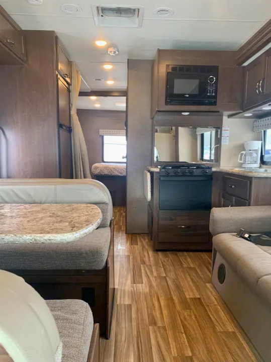 RV rental interior: kitchen with stove & microwave, dinette, and a bedroom, featuring wood floors & cabinetry.. Thor Motor Coach Four Winds 2019