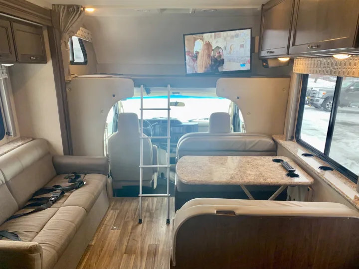 View inside an RV rental, featuring a lounge sofa with seatbelts, dining booth, TV, and ladder to the overhead bunk.. Thor Motor Coach Four Winds 2019