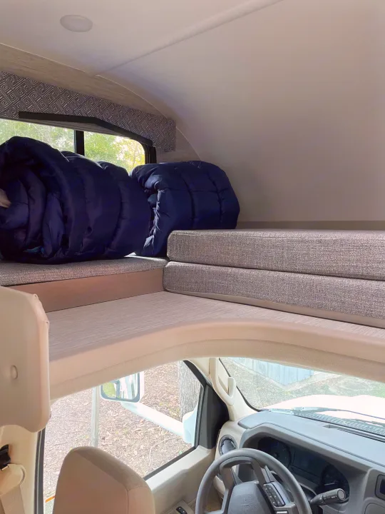 Cab-over bunk of an RV rental, seen from the driver's area. Features comfortable gray cushions & dark blue bedding.. Thor Magnitude GA22 2022