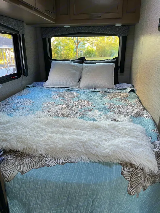 Experience comfort in this cozy RV rental bedroom. Features a blue floral bedspread, plush white throw, and window views.. Thor Magnitude GA22 2022