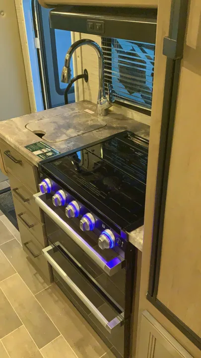Modern RV rental kitchen with a sleek stove featuring blue-lit knobs, oven, and sink. Compact design perfect for travel.. Thor Magnitude GA22 2022