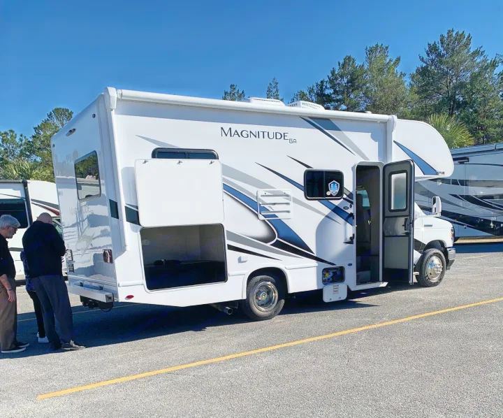 White Magnitude GA motorhome with open compartments and entry door. Two men examine this potential RV rental on a sunny day.. Thor Magnitude GA22 2022