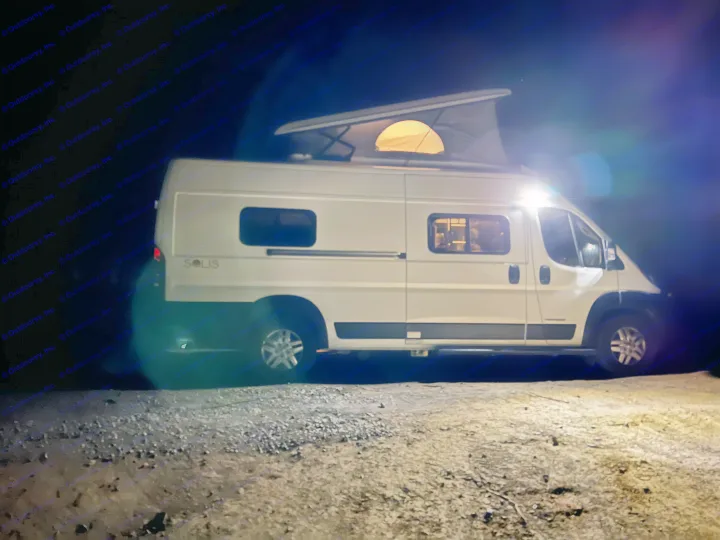 White RV rental camper van at night, with a lit pop-top tent and interior, parked on a gravel surface.. Winnebago solis 2022