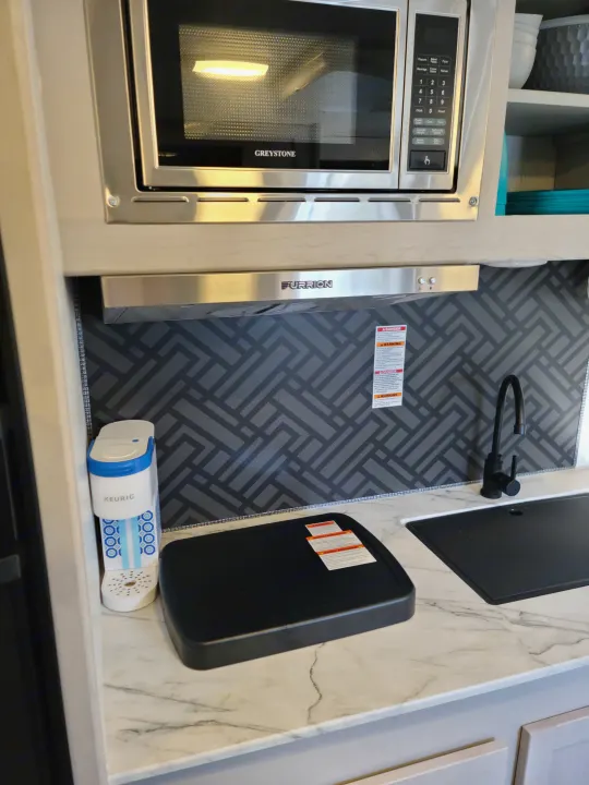 Compact RV rental kitchen with Greystone microwave, Furrion range hood, sink, and Keurig on a marble-patterned counter.. Heartland mallard 2022