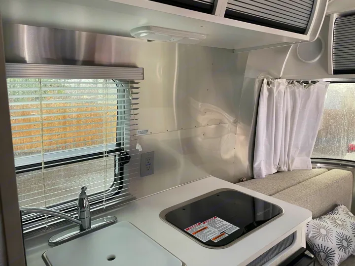 Cozy RV rental interior with a compact kitchen (sink, cooktop), window blinds, and comfortable upholstered seating.. Airstream Bambi 2021