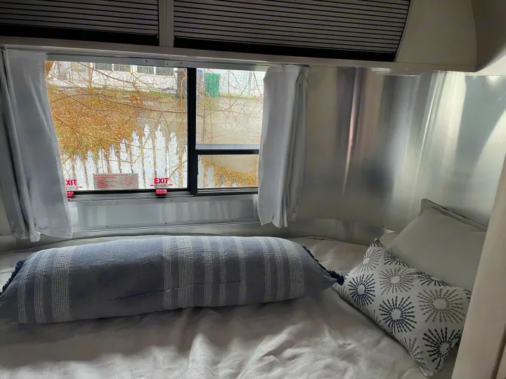 Inside an RV rental: a comfy Airstream bedroom with metallic walls, bed, striped pillow, and window view with emergency exits.. Airstream Bambi 2021